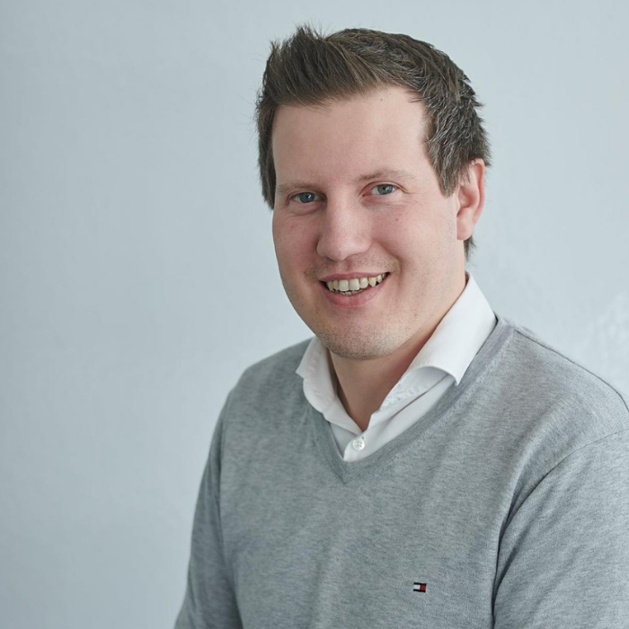 Porträt eines lächelnden Mannes in grauem Pullover und weißem Hemd vor hellem Hintergrund.