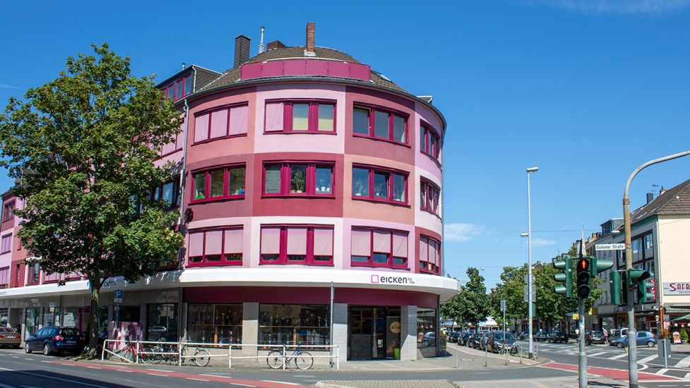 Rundes Gebäude mit pink-roter Fassade an einer Straßenecke, im Erdgeschoss Geschäfte und Bäume im Vordergrund.