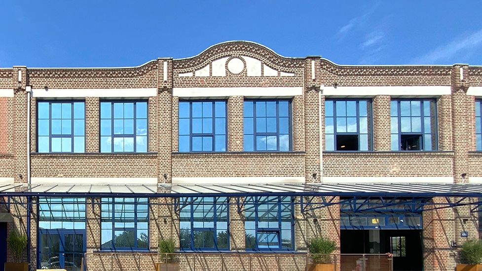 Backsteingebäude mit großen blauen Fenstern und modernem Eingangsbereich bei Sonnenschein.