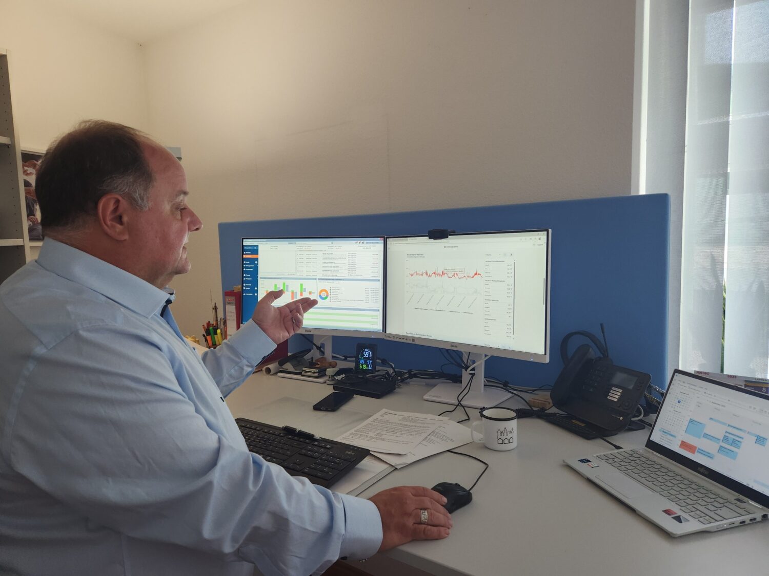 Mann im Büro arbeitet an zwei Monitoren mit Diagrammen und Statistiken, daneben ein Laptop mit geöffnetem Kalender.